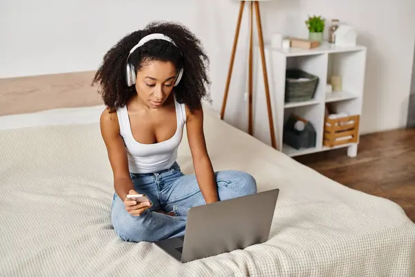 A curly African American woman in a tank top sits on a bed, using a laptop computer in a modern bedroom.