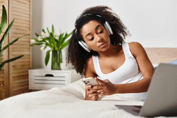 A curly African American woman in a tank top relaxes in bed, listening to music on her phone in a modern bedroom.