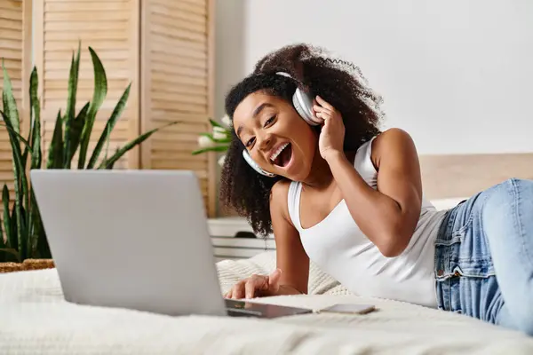 Curly African American woman relaxes on a bed with a laptop and headphones in a modern bedroom.