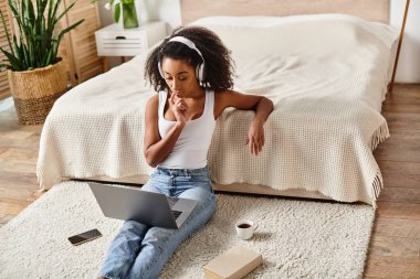 Askılı tişörtlü kıvırcık Afrikalı Amerikalı bir kadın modern yatak odası ortamında dizüstü bilgisayar kullanarak yerde oturuyor..