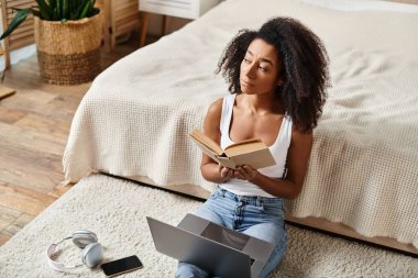 Askılı tişörtlü kıvırcık Afrikalı Amerikalı bir kadın modern bir yatak odasında yerde oturmuş kitap okumakla meşgul..
