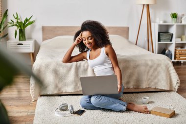 Askılı tişörtlü kıvırcık Afrikalı Amerikalı bir kadın modern bir yatak odasında dizüstü bilgisayar kullanmaya dalmış bir şekilde yerde oturuyor..
