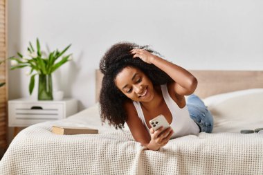 Kolsuz tişörtlü kıvırcık Afrikalı Amerikalı bir kadın cep telefonu ekranına sarılmış bir şekilde bir yatakta yatıyor..