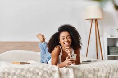 Askılı tişörtlü kıvırcık Afrikalı Amerikalı bir kadın modern bir yatak odasında elinde cep telefonu olan bir yatağa uzanır..