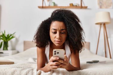 Kolsuz tişörtlü kıvırcık Afrikalı Amerikalı kadın, bir yatağa uzanmış, cep telefonuyla modern bir yatak odasına dalmış..