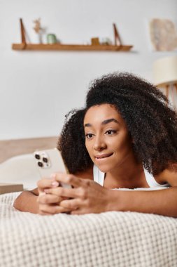 Kolsuz tişörtlü kıvırcık Afrikalı Amerikalı kadın, bir yatağa uzanmış, cep telefonuyla modern bir yatak odasına dalmış..