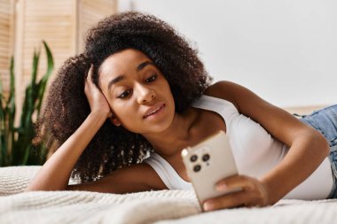 Kolsuz tişörtlü kıvırcık Afrikalı Amerikalı bir kadın. Yatakta dinleniyor, cep telefonunu modern bir yatak odasında tutuyor..