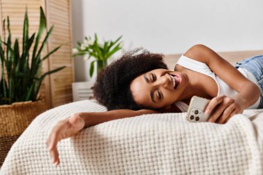 Kolsuz tişörtlü kıvırcık Afrikalı Amerikalı kadın. Yatakta dinleniyor, elinde akıllı bir telefon tutuyor..