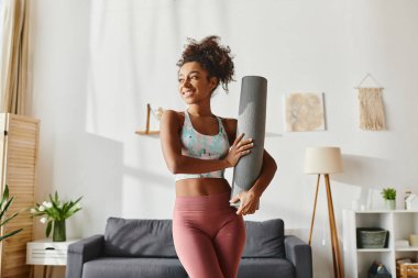 Kıvırcık Afrikalı Amerikalı etekli bir kadın şık bir oturma odasında yoga minderi tutuyor. Egzersiz için hazır..