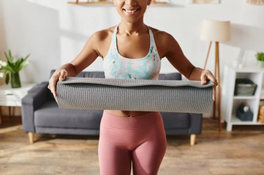 Aktif kıyafetli Afrikalı Amerikalı bir kadın bir yoga minderi tutuyor. Parlak bir gülümseme, sağlıklı ve pozitifliği somutlaştırıyor..