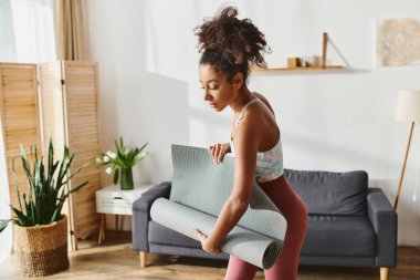 Hareketli giyimli kıvırcık saçlı Afrikalı Amerikalı kadın rahat bir oturma odasında yoga minderi tutuyor, barışçıl bir antrenmana hazır..