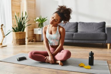 Aktif giyinmiş kıvırcık saçlı Afrikalı Amerikalı kadın yoga minderinde oturur, rahat oturma odasında farkındalık çalışması yapar..