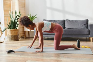 Aktif giysili kıvırcık Afrikalı Amerikalı bir kadın yoga minderinde zarif bir şekilde dengede duruyor..