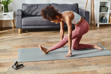 Aktif kıyafetli kıvırcık Afrikalı Amerikalı bir kadın evinde yoga minderinde yoga pozu veriyor..