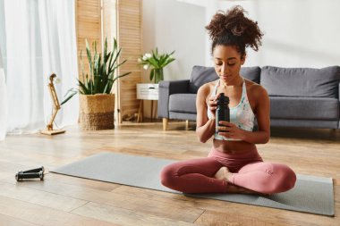 Aktif kıyafetli kıvırcık Afrikalı Amerikalı kadın yoga minderinde oturmuş, elinde bir şişe su tutuyordu..