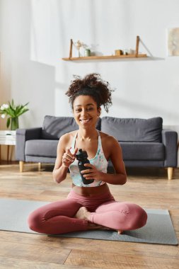 Aktif kıyafetli kıvırcık Afrikalı Amerikalı bir kadın yoga minderinde elinde bir şişe suyla oturuyor..