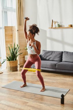 A curly African American woman practicing in living room with elastics, dressed in active wear. clipart