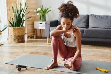 Hareketli kıvırcık Afrikalı Amerikalı bir kadın rahat bir oturma odasında yoga minderinde oturup yoga pozu veriyor..