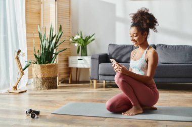 Aktif giyimli Afrikalı Amerikalı bir kadın yoga minderinde oturuyor, telefon ekranına odaklanmış..