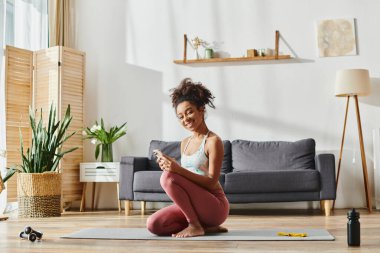 Aktif kıyafetli kıvırcık Afrikalı Amerikalı bir kadın yoga minderinde oturuyor, evde çalışırken kendini telefon ekranına kaptırmış..