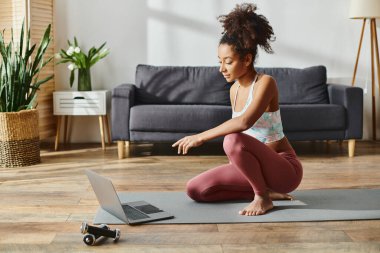 Aktif giyinmiş kıvırcık saçlı Afrikalı Amerikalı bir kadın yoga minderinin üzerinde oturuyor, kendini dizüstü bilgisayarına kaptırmış..