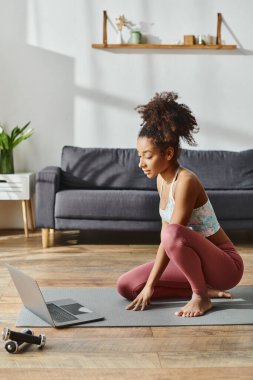 Aktif giyinmiş kıvırcık saçlı Afrikalı Amerikalı kadın yoga minderinde dizüstü bilgisayarla oturuyor ve online fitness seansına katılıyor..