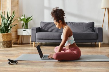 Aktif durumda olan kıvırcık saçlı Afrikalı Amerikalı kadın yoga minderinde oturup laptopunu iş için ya da dinlenmek için kullanıyor..