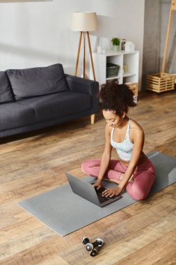 Aktif giyinmiş kıvırcık saçlı bir Afrikalı Amerikalı kadın yoga yaparken dizüstü bilgisayarına odaklanmış yoga minderinin üzerinde oturuyor..