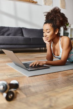 Aktif giyimde olan kıvırcık Afrikalı Amerikalı kadın dizüstü bilgisayar kullanıyor, yoga minderine uzanıyor, teknolojiyi fiziksel aktivitelerle harmanlıyor..
