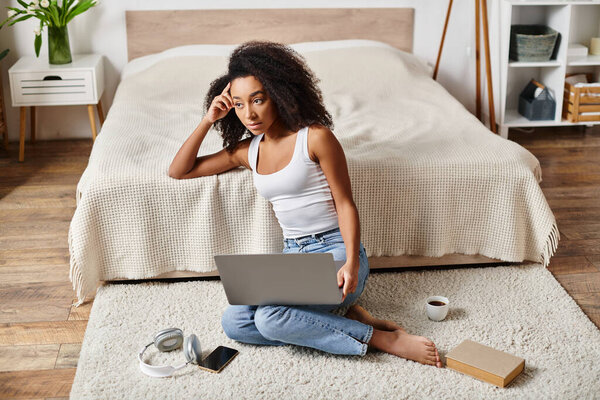 Кудрявая афроамериканка в майке сидит на полу, погруженная в использование ноутбука в современной спальне.
