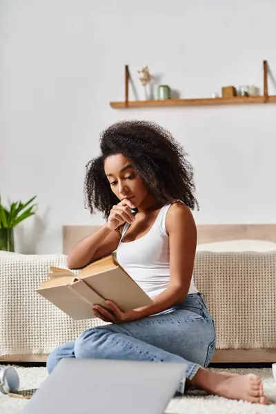 Curly African American woman in a tank top sits on the floor engrossed in a book, finding peace in her quiet moment.