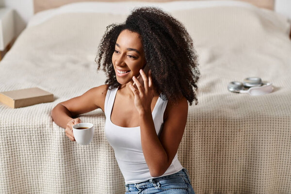 Curly African American woman in tank top enjoys a moment of relaxation, sipping coffee on a cozy bed in a modern bedroom.