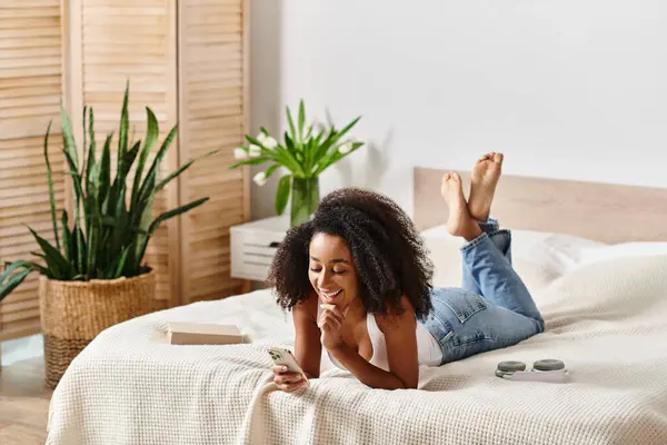 タンクトップのカーリーアフリカ系アメリカ人女性が 現代の寝室で携帯電話の画面に吸収され ベッドの上に横たわっています — ストック写真