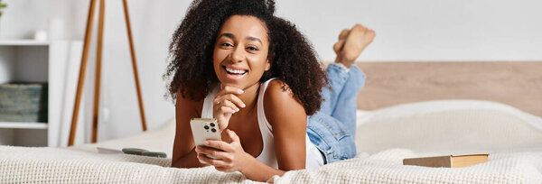 A curly African American woman peacefully lays on a bed, holding a remote in her hand in a modern bedroom.