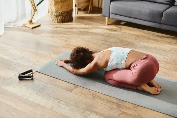 stock image A curly African American woman in activewear peacefully practices yoga on a mat.