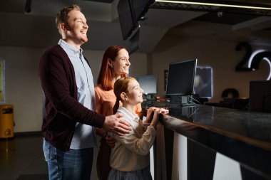 A happy man, woman, and child standing at a concession counter in a cinema, choosing snacks before enjoying a movie together. clipart