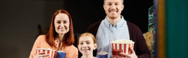 A man, woman, and child happily hold popcorn buckets at the cinema, enjoying a family movie night out. clipart