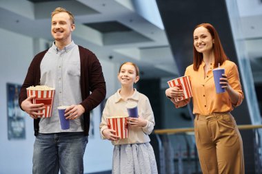 A happy family, consisting of a man, woman, and child, joyfully holding popcorn while spending quality time together at the cinema. clipart