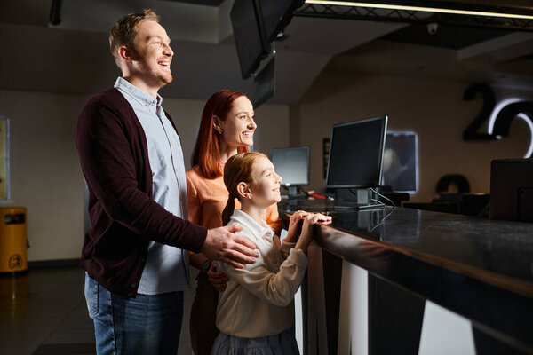 Счастливый мужчина, женщина и ребенок, стоящие у стойки концессии в кинотеатре, выбирающие закуски, прежде чем наслаждаться фильмом вместе.