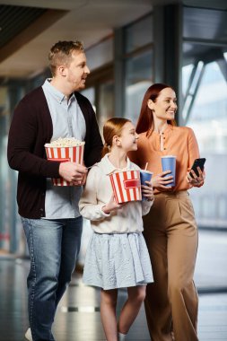 A man, woman, and child happily hold popcorn boxes in a cinema, ready to enjoy a movie together. clipart