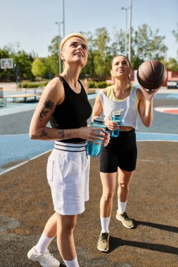 Bir basketbol sahasında kendinden emin iki genç kadın güneşli bir günde spor kıyafetleri içinde güç ve kararlılıkla ayakta duruyor..
