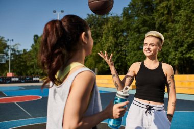 A young woman stands confidently in front of a basketball on a sunny outdoor court. clipart