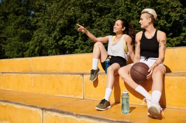 Two young women, athletic friends, sit closely together after playing basketball outdoors on a summer day. clipart