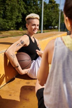 Spor kıyafetleri içindeki genç bir kadın bir bankta oturur, dışarıda huzurlu bir ifadeyle basketbol topunu beşiğinde sallar..