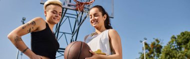 Güneşli bir günde, iki genç kadın bir basketbol topunu tutarak, açık havada oyun oynuyorlar..