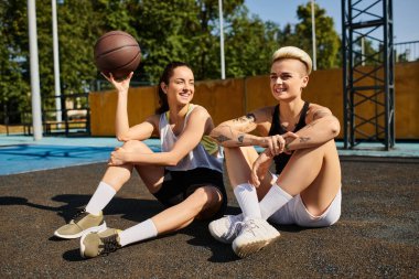 İki genç kadın, atletik ve hevesli, aralarında bir basketbol topuyla yerde oturuyor, güneşli bir yaz gününün tadını çıkarıyorlar..