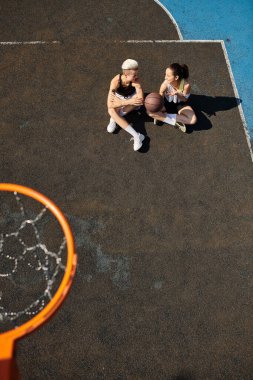 İki genç kadın basketbol sahasının tepesinde oturmuş sohbet ediyor ve yaz gününün tadını çıkarıyorlar..