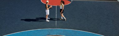 İki atletik kadın, yaz güneşinin altında yarışmaya hazır bir tenis kortunda güvenle duruyorlar..