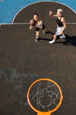 Genç bir kadın sahada basketbol oynuyor. Güneşli gökyüzünün altında top sürüyor ve basket oynuyor..