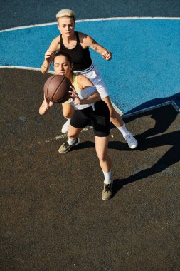 İki genç kadın, atletik ve rekabetçi, yaz güneşinin altında arkadaşça bir basketbol oyunu oynuyorlar..